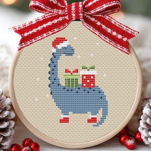Op de afbeelding: Een kruissteek kerstversiering met een blauwe dinosaurus met een kerstmuts en twee ingepakte cadeaus. Het ontwerp is geplaatst in een houten hoepel en afgewerkt met een rood-wit gestreepte strik.