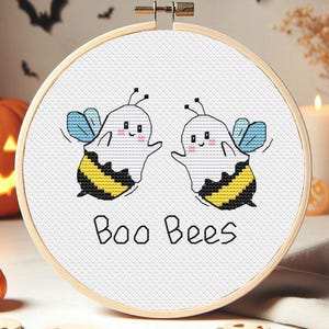 May include: A white embroidery hoop with a white background and two ghost bees with black and yellow stripes. The text "Boo Bees" is written in black below the bees.