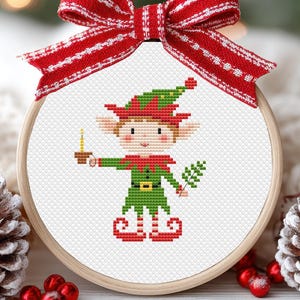 May include: A cross-stitch pattern of a Christmas elf holding a candle and a sprig of holly. The elf is wearing a green and red suit with a pointed hat. The background is white.