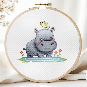 Può includere: Un telaio da ricamo a punto croce con un ippopotamo grigio con guance rosa e un piccolo uccello giallo appollaiato sulla sua testa. L'ippopotamo è in acqua con fiori rosa. Il ricamo è in un telaio di legno rotondo.