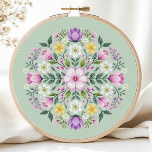 May include: A circular embroidery hoop with a green background and a floral design. The design features pink, white, and purple flowers with green leaves.