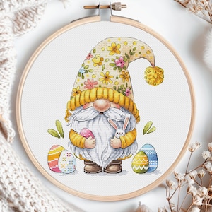 Op de afbeelding: Een gele en bloemige gnome die een wit konijn en twee kleurrijke paaseieren vasthoudt. De gnome draagt ​​een gele hoed met een bloemenpatroon en een witte baard. De gnome staat op een witte achtergrond.