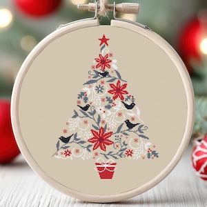 May include: A cross-stitch embroidery hoop with a beige background featuring a stylized Christmas tree design. The tree is decorated with red poinsettias, white snowflakes, and small gray birds. A red gift box sits at the base of the tree.