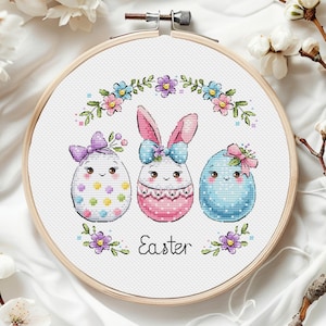 Puede incluir: Un aro de bordado de punto de cruz con fondo blanco que presenta tres huevos de Pascua. Los huevos están decorados con lunares coloridos, flores y lazos. El huevo del centro tiene orejas de conejo y un lazo rosa. El texto "Easter" está escrito debajo de los huevos.