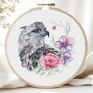 Może przedstawiać: Wzór do haftu krzyżykowego przedstawiający szczegółową ilustrację jastrzębia z szaro-czarnym upierzeniem, otoczonego różowymi i fioletowymi kwiatami. Wzór jest oprawiony w drewniany tamborek do haftu.