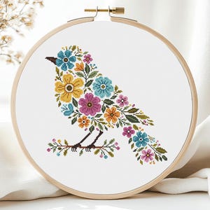 May include: A circular wooden embroidery hoop containing a cross-stitch pattern of a bird silhouette formed by various colorful flowers. The bird features blue, yellow, orange, and pink blossoms with green leaves, perched on a brown branch.