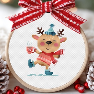 May include: A cross-stitch embroidery hoop featuring a cartoon reindeer wearing a red scarf, hat, and ice skates, holding a mug. The reindeer is smiling and the hoop is decorated with a red and white striped bow. The embroidery is set against a white background.