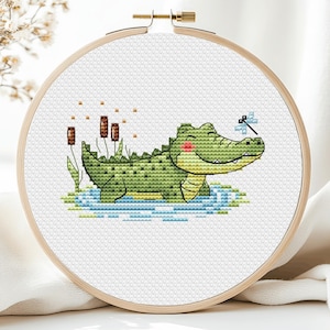 May include: Cross-stitch embroidery in a wooden hoop features a smiling green alligator in water with a dragonfly. The alligator has a pink blush and is surrounded by cattails and green plants. The background is white.