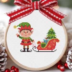 May include: A cross-stitch Christmas ornament featuring a cartoon elf with a red and green outfit, pulling a sleigh filled with gifts and a Christmas tree. The ornament is framed in a wooden hoop and adorned with a red and white striped bow.