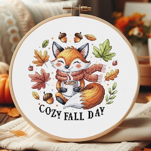Könnte beinhalten: Ein Kreuzstich-Stickrahmen mit einem Cartoon-Fuchs mit orangefarbenem und weißem Fell, der einen roten Schal trägt und eine Tasse hält. Das Design umfasst Herbstblätter, Eicheln und den Text "Cozy Fall Day."