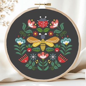 May include: A completed cross-stitch embroidery in a wooden hoop. The design features a symmetrical arrangement of flowers, a bee, and a ladybug in red, yellow, blue, and green on a dark gray background. The hoop is secured with a brass screw.