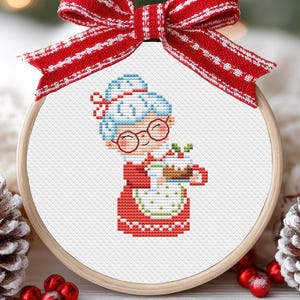 May include: A cross-stitch embroidery hoop featuring a festive design. The artwork depicts a smiling, bespectacled woman with blue hair, wearing a red dress and apron, holding a mug. A red and white striped bow adorns the top. The hoop is surrounded by Christmas decorations.