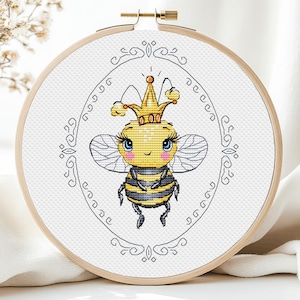 May include: A cross-stitch pattern of a cute cartoon bee wearing a gold crown. The bee is yellow with black stripes and has big blue eyes. The pattern is framed in a wooden embroidery hoop.