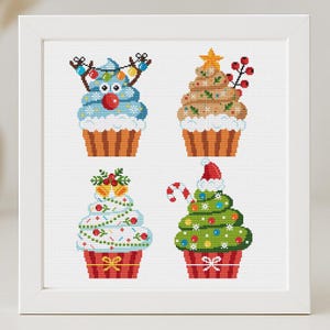 May include: Four Christmas-themed cupcakes with different decorations. One cupcake has a blue reindeer with red antlers and lights, another has a brown tree with a yellow star, a third has a white tree with green and white decorations, and the last has a green tree with a red Santa hat.