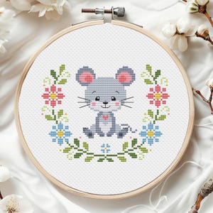 Peut inclure: Broderie au point de croix d'une souris grise aux oreilles roses et un cœur, entourée d'une couronne florale dans un cercle en bois. La couronne présente des fleurs et des feuilles roses, bleues et vertes, créant un design charmant et fait à la main.