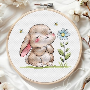 May include: A cross-stitch embroidery pattern featuring a brown bunny with pink ears, smiling and holding its paws together, in front of a blue flower with green leaves. Two yellow bees are flying above the bunny.