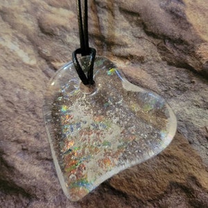 May include: Clear heart-shaped pendant with iridescent accents, suspended from a black cord. The pendant has a textured, glittery appearance, with a hole at the top for the cord. The background is a textured, brown and gray surface.