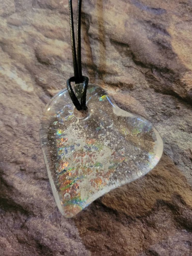 May include: Clear heart-shaped pendant with iridescent accents, suspended from a black cord. The pendant has a textured, glittery appearance, with a hole at the top for the cord. The background is a textured, brown and gray surface.