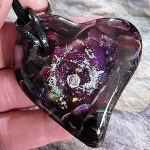 May include: A heart-shaped pendant with a dark purple and clear glass design. The pendant has a black cord and features a swirling pattern with iridescent accents. The glass has a glossy finish.