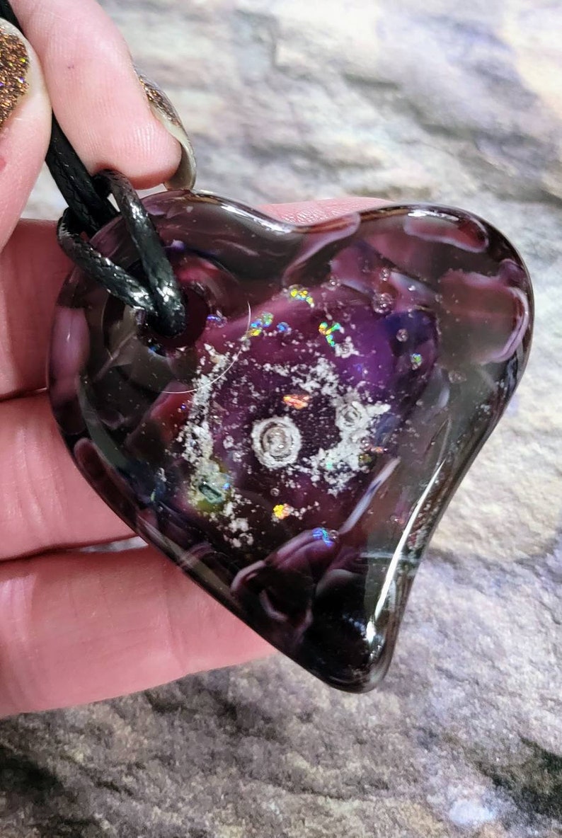May include: A heart-shaped pendant with a dark purple and clear glass design. The pendant has a black cord and features a swirling pattern with iridescent accents. The glass has a glossy finish.