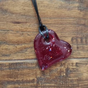 May include: A heart-shaped pendant necklace. The pendant is a deep red color with glitter accents, suspended from a black cord. The necklace is displayed on a wooden surface.