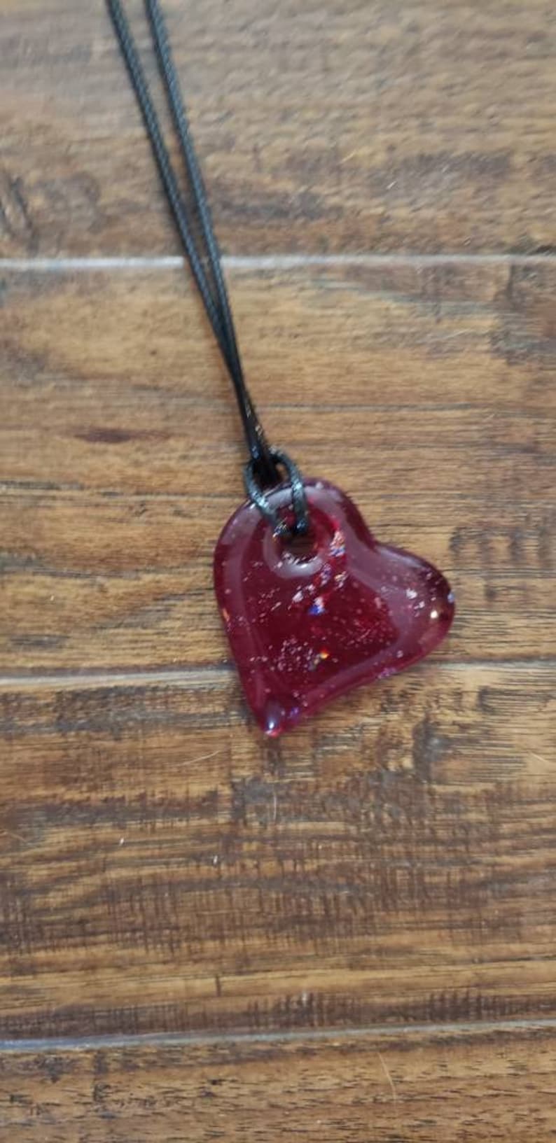 May include: A heart-shaped pendant necklace. The pendant is a deep red color with glitter accents, suspended from a black cord. The necklace is displayed on a wooden surface.