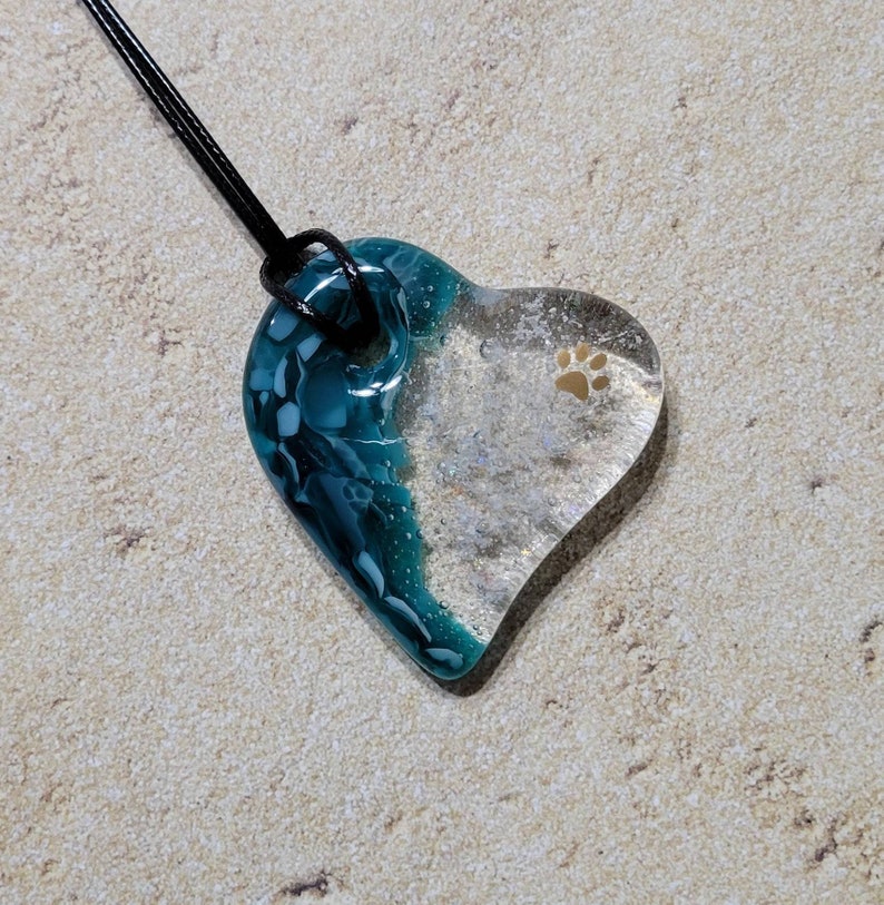 May include: Heart-shaped pendant with a black cord. The pendant is half teal and white, and half clear with a tan paw print. The pendant is made of glass and has a glossy finish.