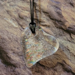 May include: Clear, heart-shaped pendant with iridescent glitter suspended within. The pendant is strung on a black cord. The heart design is visible against a textured, brown background, creating a striking contrast.