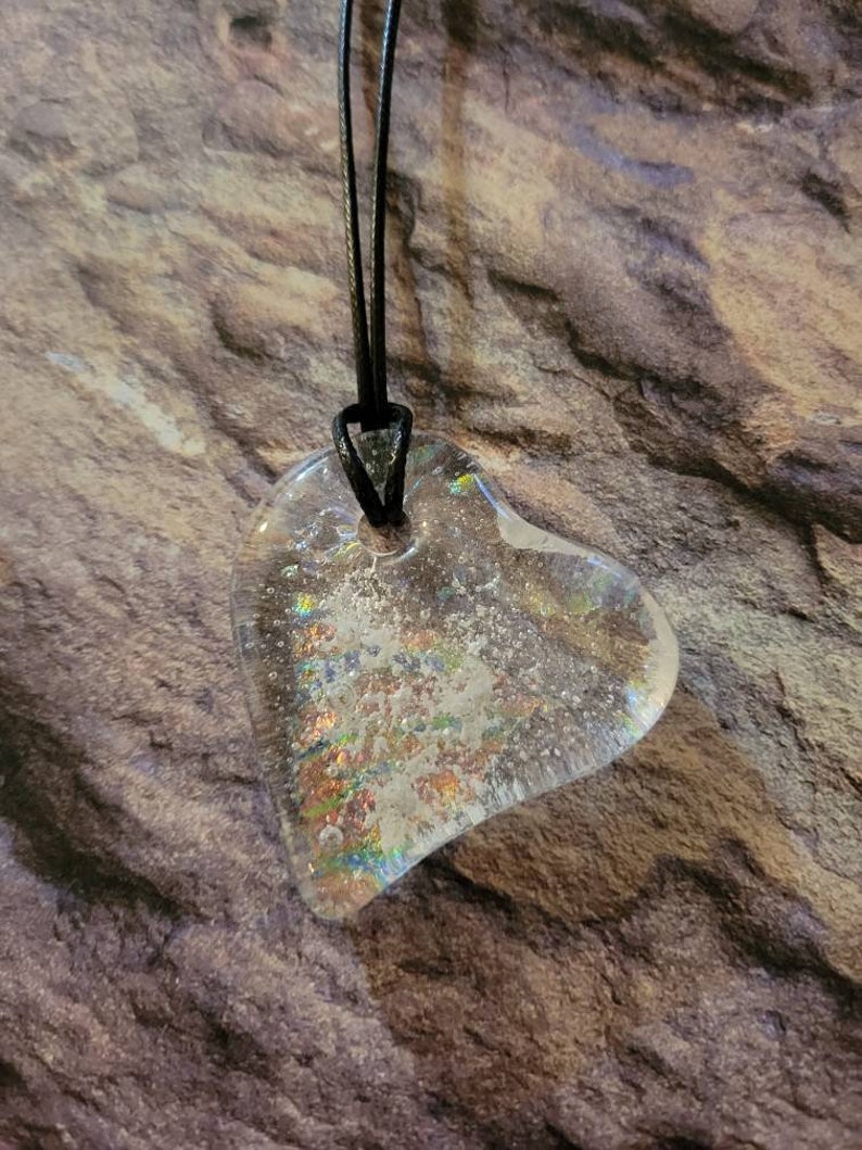 May include: Clear, heart-shaped pendant with iridescent glitter suspended within. The pendant is strung on a black cord. The heart design is visible against a textured, brown background, creating a striking contrast.