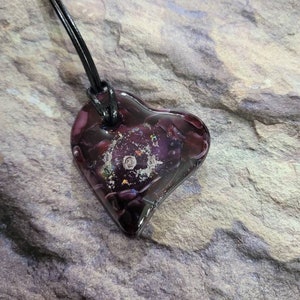 May include: A heart-shaped pendant with a dark purple hue, suspended from a black cord. The pendant has a textured, galaxy-like interior with flecks of silver and other colors. The cord is approximately 18 inches long.