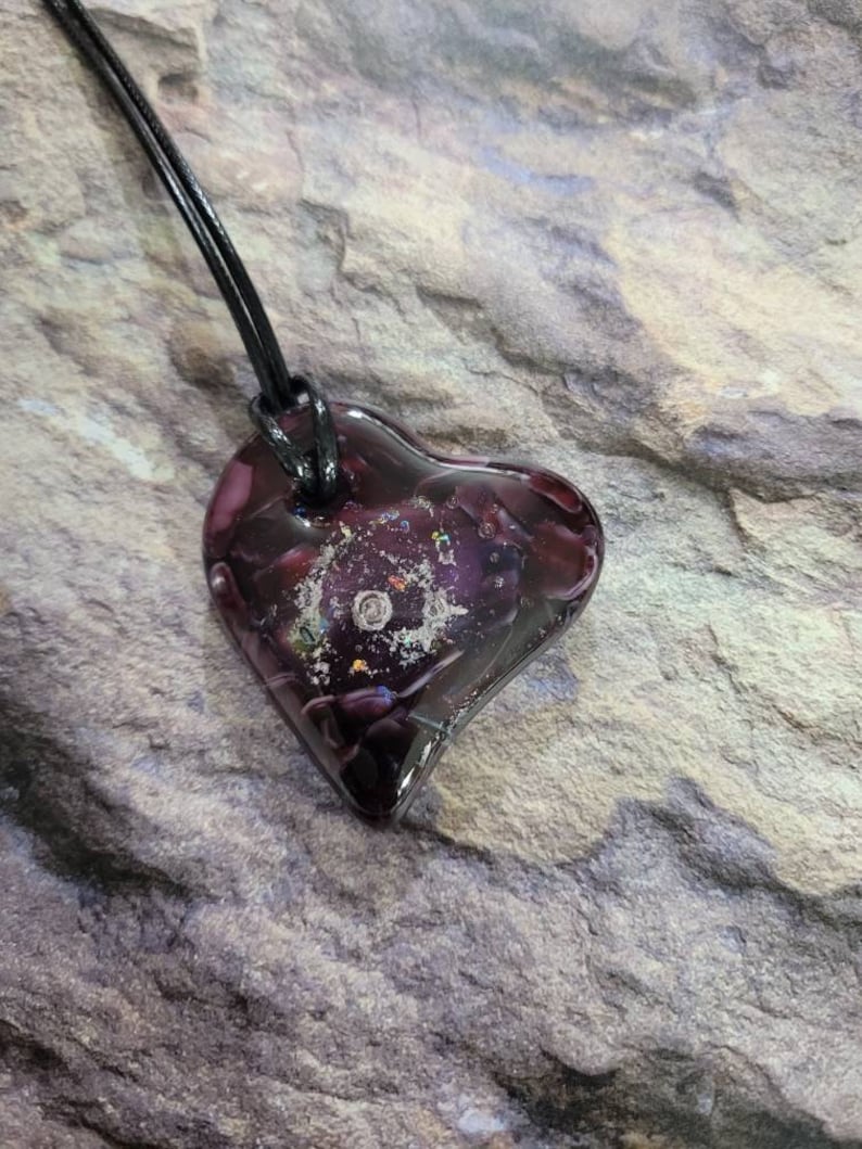 May include: A heart-shaped pendant with a dark purple hue, suspended from a black cord. The pendant has a textured, galaxy-like interior with flecks of silver and other colors. The cord is approximately 18 inches long.