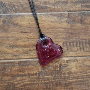 May include: A heart-shaped pendant necklace with a dark red, glitter-filled resin charm. The pendant hangs from a black cord against a wooden background. A unique piece of jewelry.