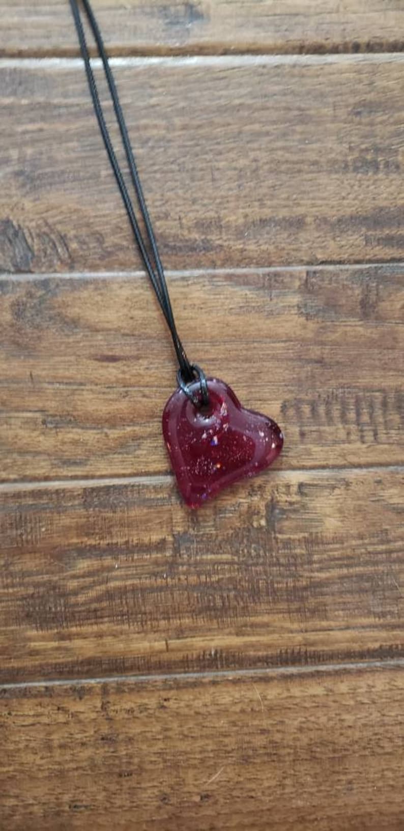 May include: A heart-shaped pendant necklace with a dark red, glitter-filled resin charm. The pendant hangs from a black cord against a wooden background. A unique piece of jewelry.