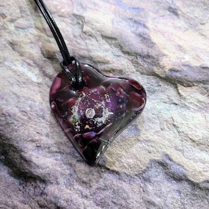 May include: A heart-shaped pendant necklace. The pendant is a deep purple color with a galaxy-like design inside. The necklace has a black cord. The pendant is approximately 1.5 inches wide and 1.5 inches tall.