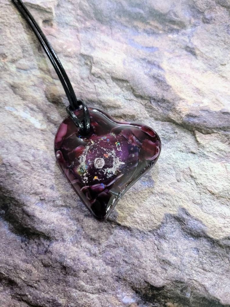 May include: A heart-shaped pendant necklace. The pendant is a deep purple color with a galaxy-like design inside. The necklace has a black cord. The pendant is approximately 1.5 inches wide and 1.5 inches tall.