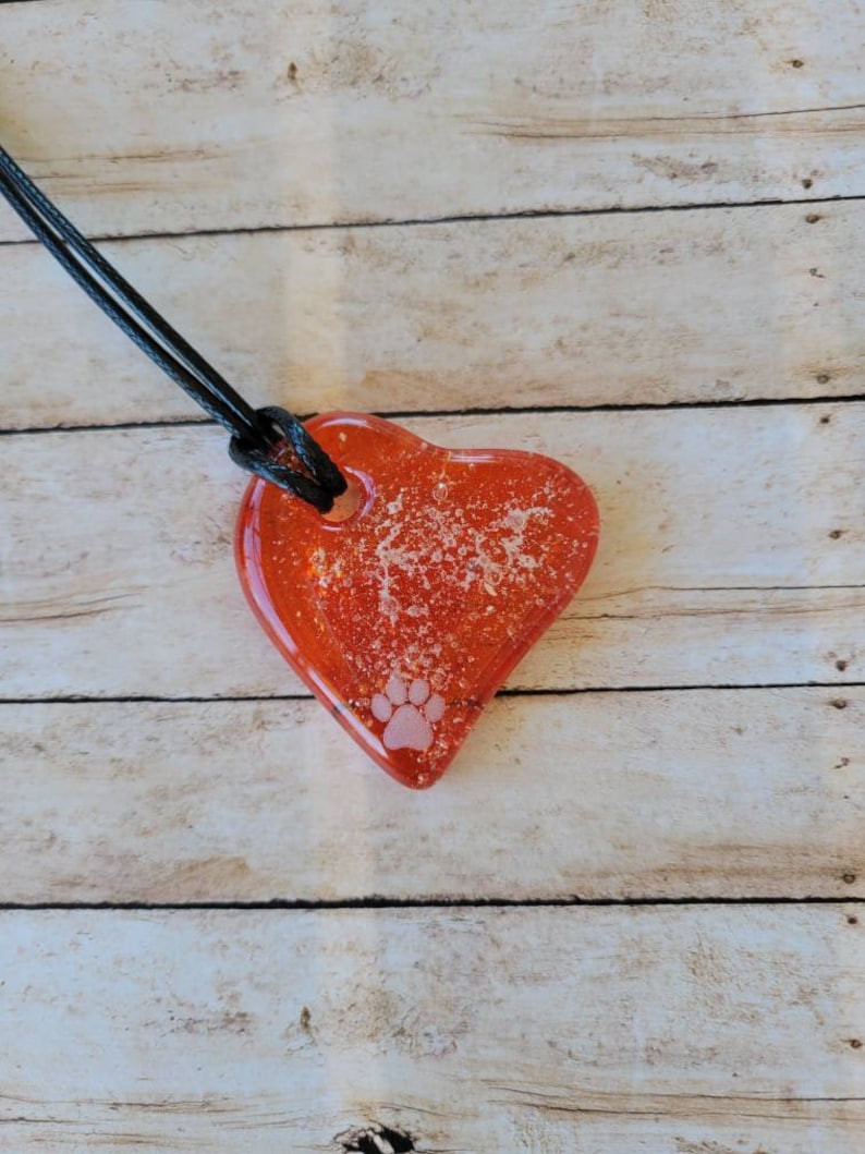 May include: A heart-shaped pendant necklace with a red, translucent resin heart. The heart has a paw print and glitter embedded within. The necklace has a black cord.