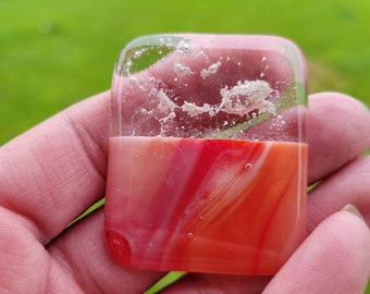 May include: A square, clear glass pendant with a red, white, and orange swirl design inside. The glass is clear and has small air bubbles trapped inside.