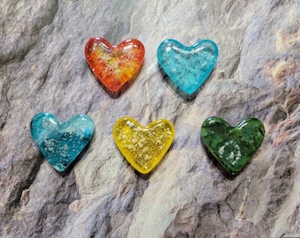 May include: Five heart-shaped glass ornaments in shades of red, blue, yellow, and green. Each heart has a unique internal design, with some appearing to have embedded elements. The ornaments are displayed on a textured, gray surface.