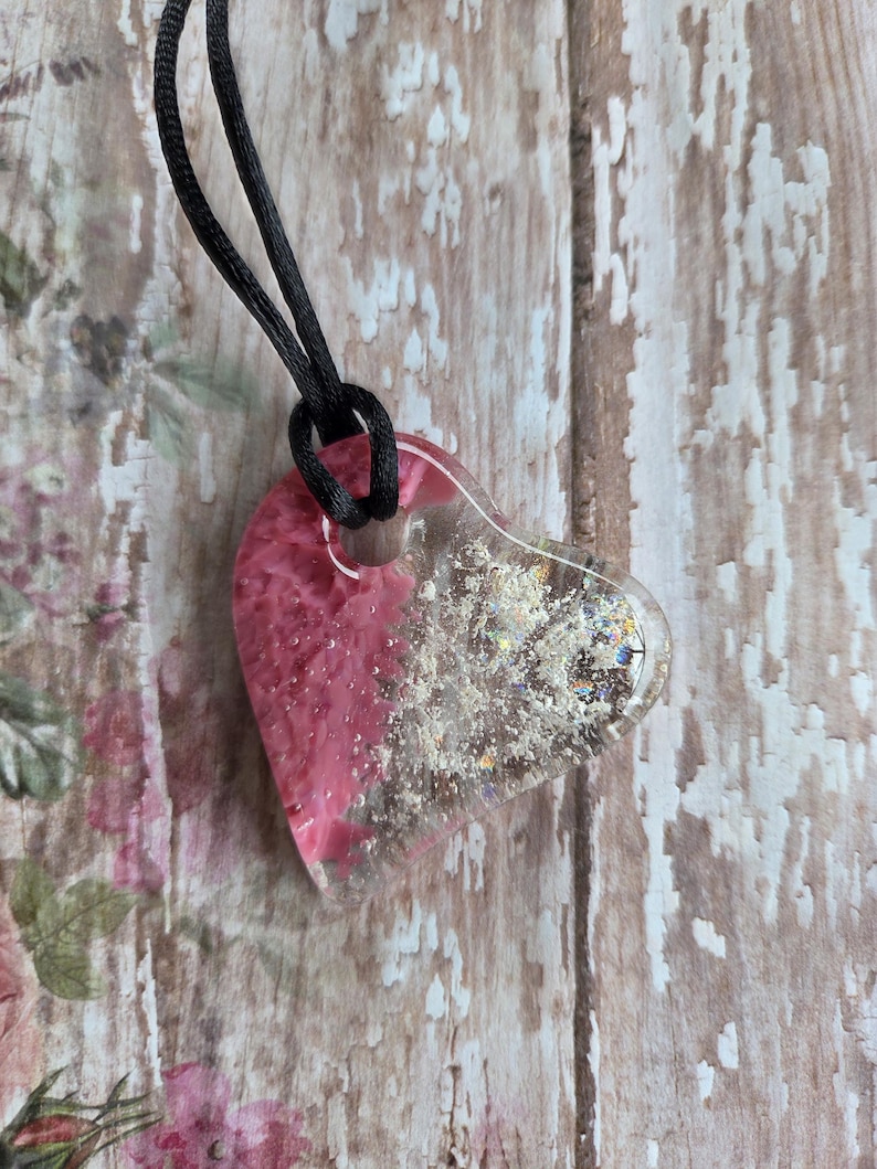 May include: A heart-shaped pendant necklace with a black cord. The pendant is clear resin with pink and silver glitter accents. The pink resin fills the left side of the heart, while the right side is clear with glitter.