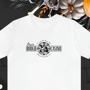 Pu&ograve; includere: T-shirt bianca con grafica nera che recita "Oogie Boogie Casino" con un design circolare e il testo "I'm a gambler, Boogie Man, although I don't play fair!". Abbigliamento a tema Halloween.