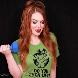 K&ouml;nnte beinhalten: Hellgr&uuml;nes T-Shirt mit einer Cartoon-Grafik einer Figur und dem Text "DO YOU EVEN LIFT?". Eine blaue Hantel wird in der Hand gehalten. Die Person l&auml;chelt. Der Hintergrund ist schwarz.