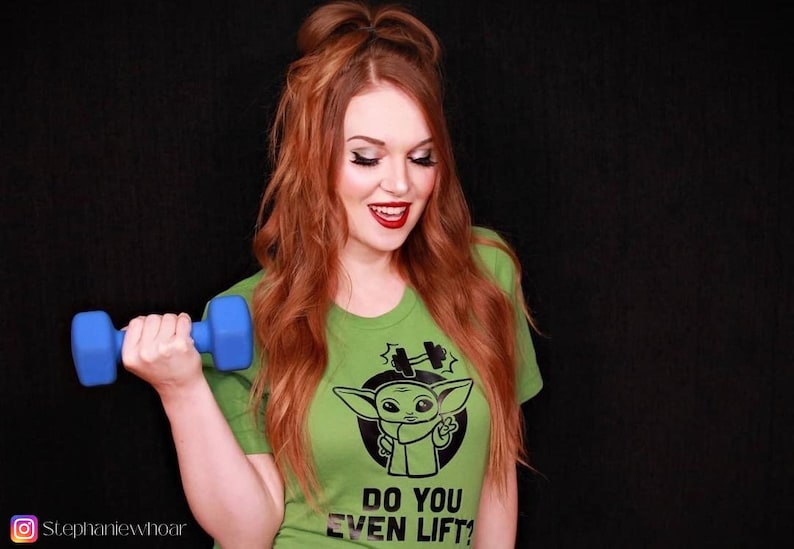 K&ouml;nnte beinhalten: Hellgr&uuml;nes T-Shirt mit einer Cartoon-Grafik einer Figur und dem Text "DO YOU EVEN LIFT?". Eine blaue Hantel wird in der Hand gehalten. Die Person l&auml;chelt. Der Hintergrund ist schwarz.