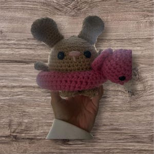 May include: A tan crocheted plush toy shaped like a rabbit with black button eyes and a small pink nose. The rabbit is wearing a bright pink crocheted inner tube floatie around its middle, which features a small pink flamingo head attached.