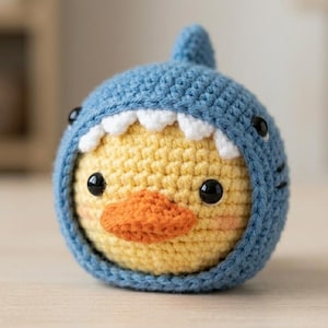 May include: A round, crocheted plush toy featuring a yellow duckling face with black eyes and an orange beak, encased in a blue shark hood with a dorsal fin, white triangular teeth, and small black side fins, crafted with visible yarn stitches.