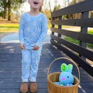 May include: A child wearing light blue pajamas with a white bunny pattern. The child is standing on a wooden bridge next to a brown basket containing a blue stuffed bunny with green ears. The child is wearing brown slippers.