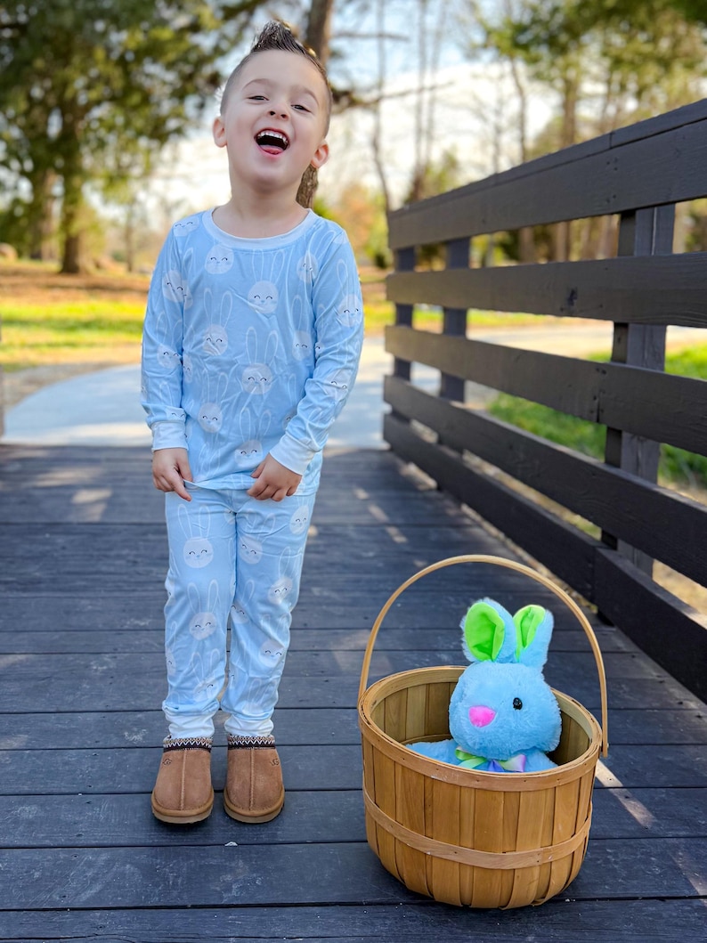 May include: A child wearing light blue pajamas with a white bunny pattern. The child is standing on a wooden bridge next to a brown basket containing a blue stuffed bunny with green ears. The child is wearing brown slippers.