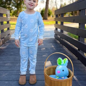 May include: A child wearing light blue pajamas with a white bunny pattern. The child is standing on a wooden bridge next to a basket containing a blue stuffed bunny with green ears. The child is wearing brown boots.