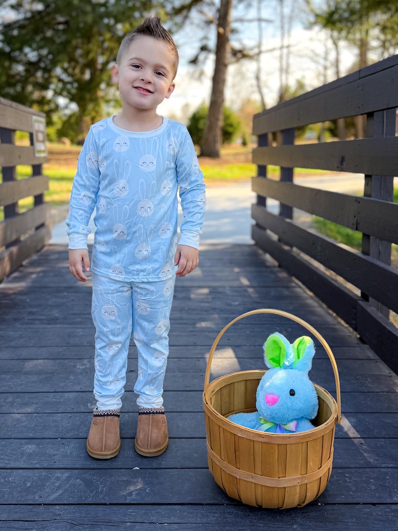 May include: A child wearing light blue pajamas with a white bunny pattern. The child is standing on a wooden bridge next to a basket containing a blue stuffed bunny with green ears. The child is wearing brown boots.