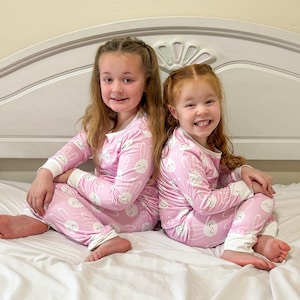 May include: Two young girls wearing matching pink pajamas with a white bunny pattern. The pajamas have long sleeves and pants with white cuffs. They are sitting on a white bed, smiling at the camera.