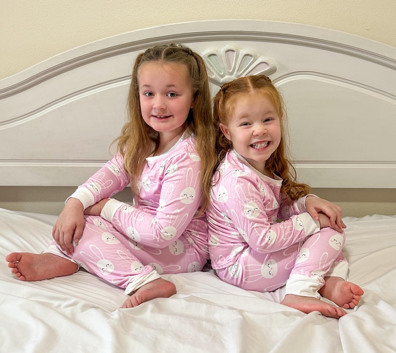 May include: Two young girls wearing matching pink pajamas with a white bunny pattern. The pajamas have long sleeves and pants with white cuffs. They are sitting on a white bed, smiling at the camera.
